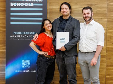Drei Personen stehen vor einem Max Planck Schools Banner.
