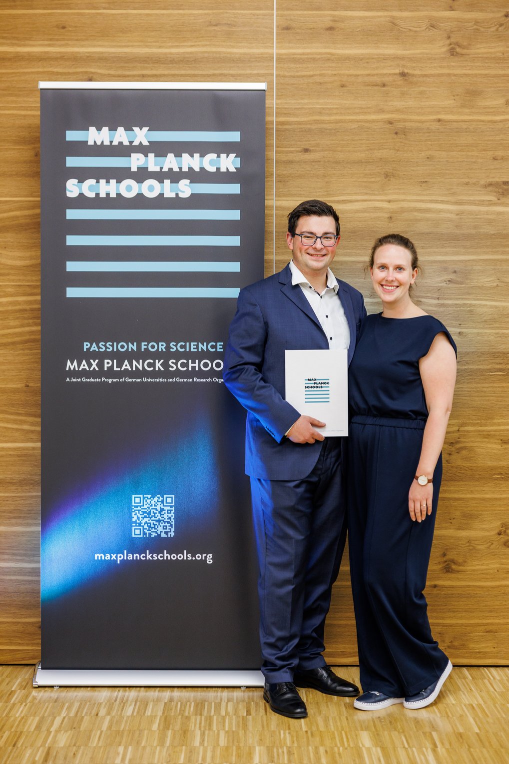 Zwei Personen stehen vor einem Banner der Max Planck Schools. Der Mann trägt einen Anzug und hält einen Flyer. Beide Personen sind elegant gekleidet. Das Banner zeigt das Logo der Max Planck Schools und einen QR-Code.