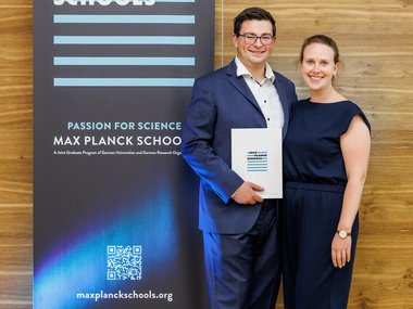 Two people are standing in front of a Max Planck Schools banner. The man is wearing a suit and holding a flyer. Both individuals are elegantly dressed. The banner displays the Max Planck Schools logo and a QR code.