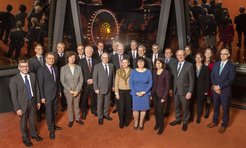 Eine Gruppe von Personen in formeller Geschäftskleidung steht in einem Raum mit großen Fenstern, durch die eine nächtliche Stadtszene mit einem hell erleuchteten Riesenrad im Freien zu sehen ist.