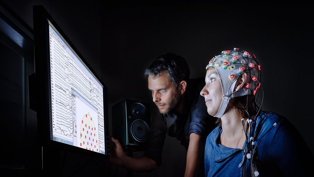 A person is seated wearing an EEG cap equipped with numerous sensors, observing brainwave data displayed on a computer monitor nearby.