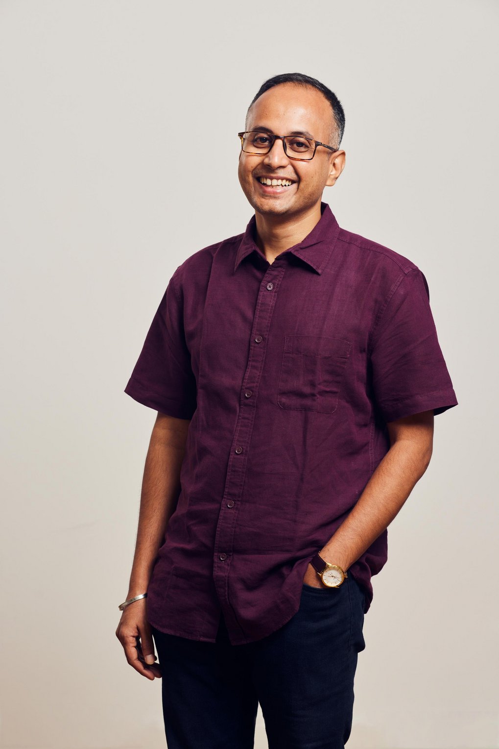 A person stands wearing a burgundy short-sleeved shirt with button details, paired with dark trousers. One hand is casually placed in a pocket, and the wrist features a watch. The background is neutral, enhancing the focus on the clothing and accessories.