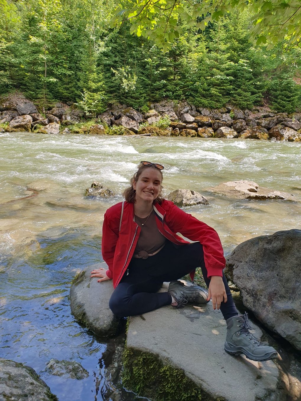 A person wearing a red jacket and hiking boots is sitting on a rock by the riverbank, surrounded by lush greenery and trees.