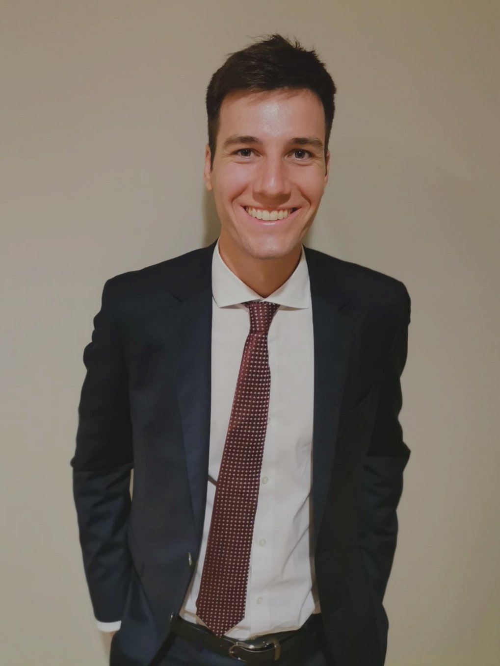 A person wearing a dark blue suit, paired with a white shirt and a red-patterned tie, stands in front of a bright background. Hands in their trouser pockets.