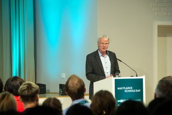 Eröffnet wurde der Tag durch eine Willkommensrede von Professor Ferdi Schüth, Mitglied des Lenkungsausschusses der Max Planck Schools. Der Chemiker ist zudem Direktor am Max-Planck-Institut für Kohlenforschung in Mülheim (Ruhr). Ein Redner steht am Pult und adressiert das Publikum beim Max Planck Schools Day. Der Raum ist in blauem Licht beleuchtet.