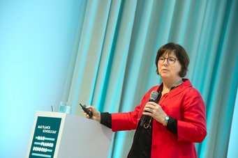 Karin Jacobs is Vice-President of the German Research Foundation (DFG) and Professor of Experimental Physics at Saarland University, with research interests in soft matter and biophysics. The person in this picture is wearing a red jacket and speaking on stage. They are holding a microphone in their right hand and a pointer in their left hand. In the background, there is a gray wall and a table with a glass of water.