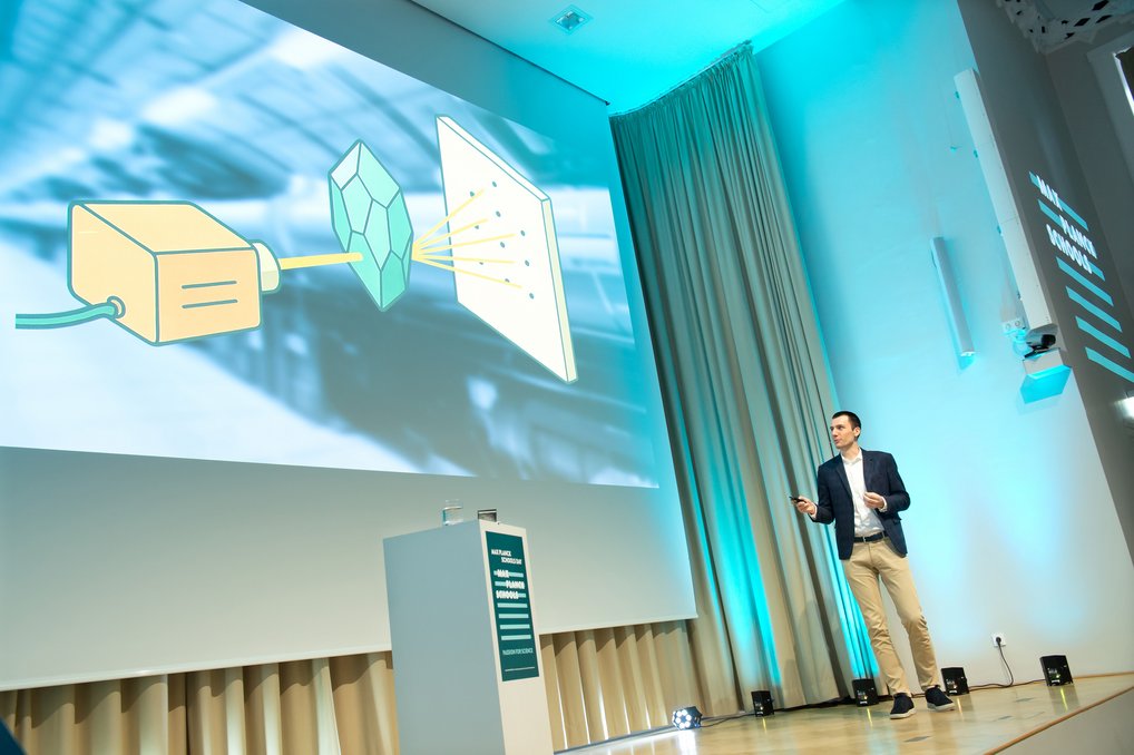 Ein Vortragender steht vor einer großen Leinwand, auf der ein Diagramm zu sehen ist, das einen Projektor, einen Kristall und eine Projektionsfläche zeigt.