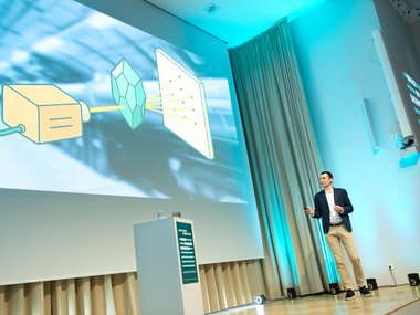 Ein Vortragender steht vor einer großen Leinwand, auf der ein Diagramm zu sehen ist, das einen Projektor, einen Kristall und eine Projektionsfläche zeigt.