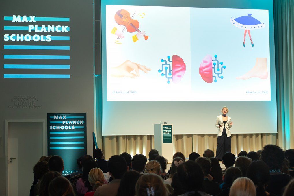 Vortragende bei den Max Planck Schools vor Publikum, Leinwand zeigt grafische Elemente und Illustrationen, Raum beleuchtet.