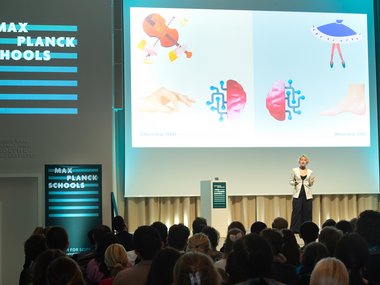 Vortragende bei den Max Planck Schools vor Publikum, Leinwand zeigt grafische Elemente und Illustrationen, Raum beleuchtet.