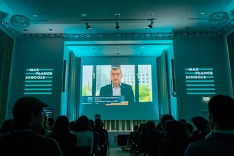 In his video message, Professor Walther Rosenthal, co-chair of the Max Planck Schools steering committee, offered encouraging words to the PhD candidates, emphasizing the importance of interdisciplinary collaboration and scientific curiosity. Audience in a dimly lit conference room, watching a presentation on a large screen with "Max Planck Schools" branding.