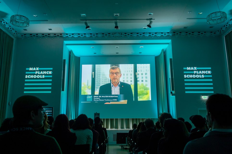 Ein abgedunkelter Raum mit Zuschauern, die einen großen Bildschirm betrachten. An den Wänden sind "Max Planck Schools"-Logos zu sehen.