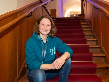A person in a teal hoodie and jeans sits on red-carpeted stairs with wooden railings, in a building with arched architecture.