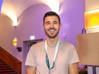 A person in a light-colored shirt and lanyard stands in a room with purple lighting, near a staircase and a lamp.