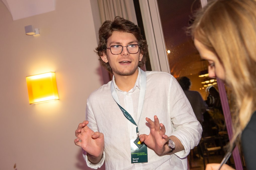 Eine Person mit weißem Hemd und Namensschild gestikuliert in einem Raum. Eine weitere Person ist teilweise sichtbar. Im Hintergrund ist ein Fenster mit Vorhängen und gelbem Wandlicht zu sehen.