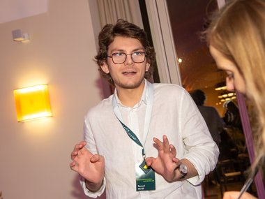 Person in a white shirt gesturing with hands, wearing a lanyard and watch, standing indoors under warm lighting.