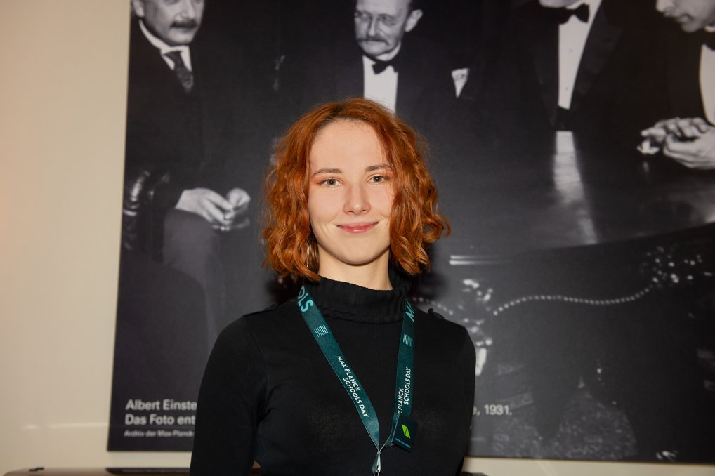 Eine Person mit roten Haaren trägt ein Namensschild und steht vor einem Schwarz-Weiß-Foto von Albert Einstein und anderen Wissenschaftlern.