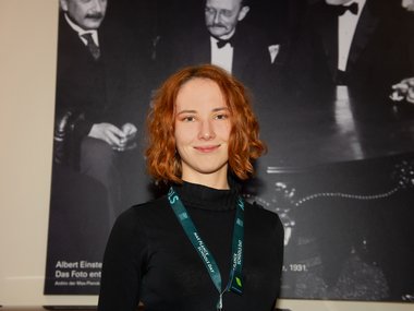A person with a green lanyard stands in front of a large black-and-white photograph, depicting Albert Einstein seated at a table with other historical figures.