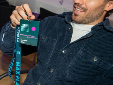 A person in a blue shirt holds a badge labeled "Max Planck School of Cognition" with the name "Conrad Blau" visible.