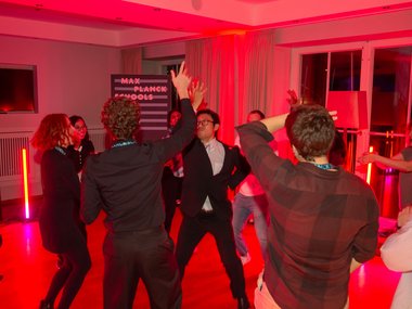 Individuals are energetically dancing in a red-lit room. The setting includes a sign labeled "Max Planck Schools," adding context to the event.