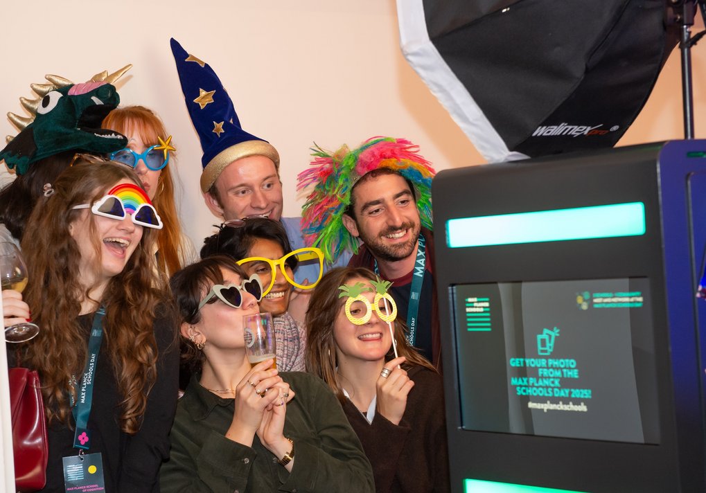 Menschen tragen bunte Hüte und Brillen, während sie vor einem Fotobox-Bildschirm stehen, der Informationen zum Max Planck Schools Day anzeigt.