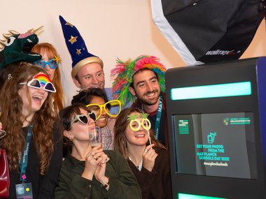 Menschen tragen bunte Hüte und Brillen, während sie vor einem Fotobox-Bildschirm stehen, der Informationen zum Max Planck Schools Day anzeigt.