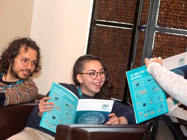 Several individuals are seated on couches, reading brochures titled "Zu den Max Planck Schools". The brochures feature educational icons and graphics.
