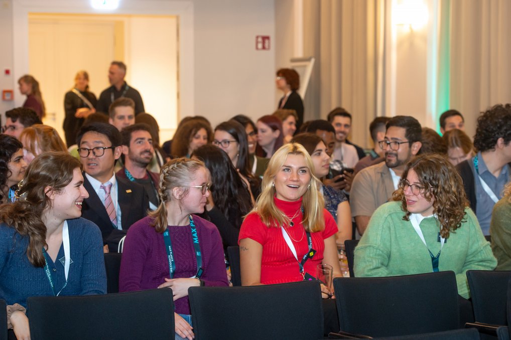 Versammlungsraum mit sitzenden Teilnehmern, viele tragen Namensschilder und Ausweise, während im Hintergrund Personen stehen.