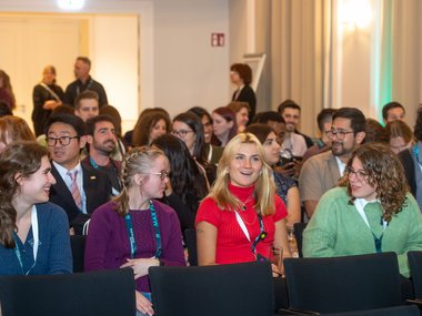A group of people in a conference room, seated in rows. Many wear conference lanyards, indicating an organized event.