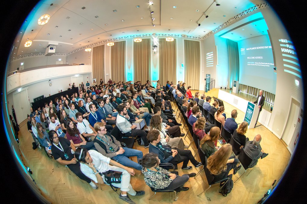 Ein gut besuchter Konferenzraum mit Reihen von Zuhörern, die einem Redner auf der Bühne aufmerksam zuhören. Der Raum ist mit hellen Lichtern und großen Vorhängen gestaltet.