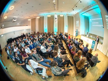 Attendees seated in a well-lit conference room, listening to a speaker on stage.