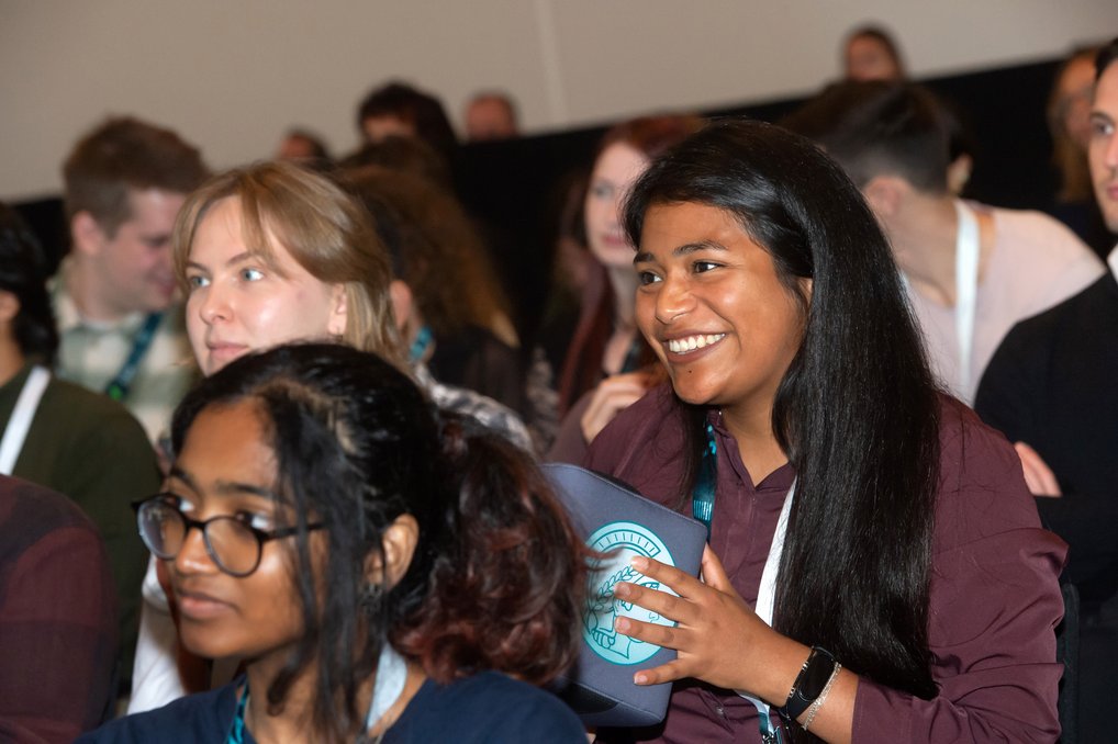 Teilnehmerin hält Mikrofonball während einer Konferenz, um Fragen zu stellen. Im Hintergrund weitere Personen mit Namensschildern.
