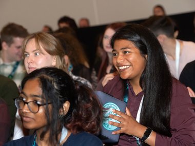 Teilnehmerin hält Mikrofonball während einer Konferenz, um Fragen zu stellen. Im Hintergrund weitere Personen mit Namensschildern.