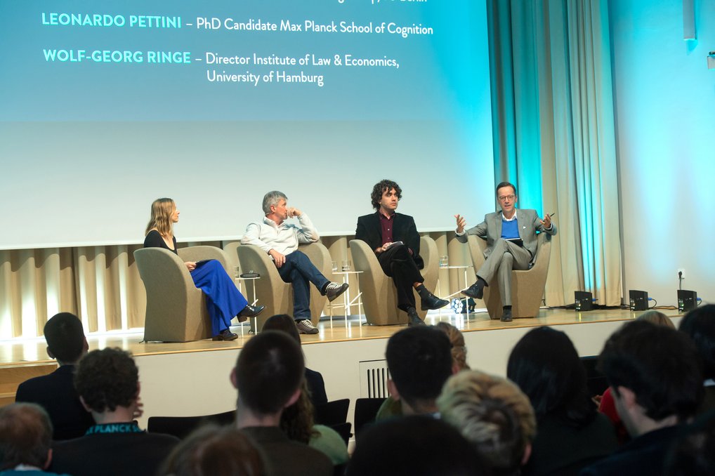 Eine Podiumsdiskussion mit vier Personen. Zwei sind namentlich auf einer Präsentation genannt. Publikum sitzt aufmerksam im Vordergrund.