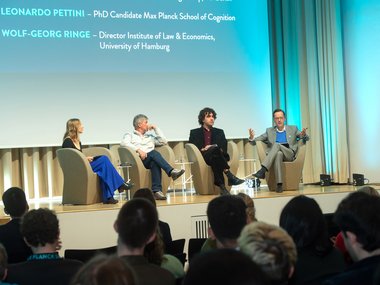 Eine Podiumsdiskussion mit vier Personen. Zwei sind namentlich auf einer Präsentation genannt. Publikum sitzt aufmerksam im Vordergrund.