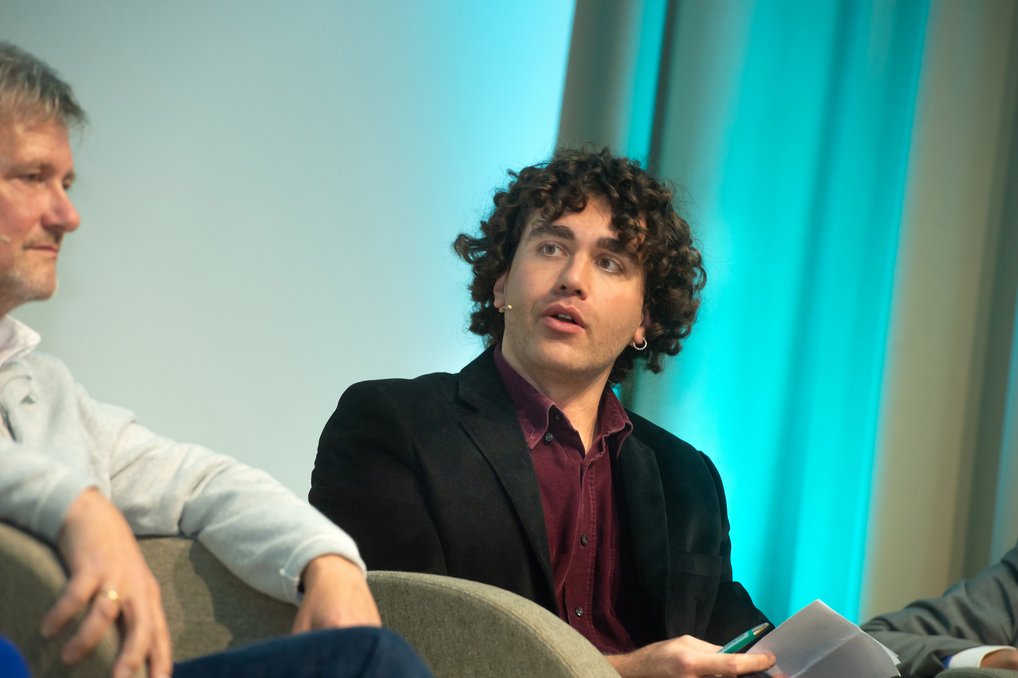 Auf einem Podium sitzt eine Person mit lockigem Haar und dunklem Sakko, die ein Notizbuch in der Hand hält. Im Hintergrund türkisfarbene Vorhänge.