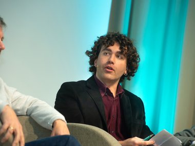 Individual in a black blazer with curly hair holds papers while seated on a panel discussion.