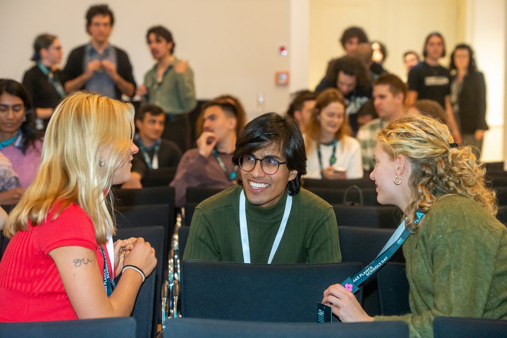Zwei Frauen und ein Mann führen ein Gespräch in einem Konferenzraum. Andere Teilnehmer stehen im Hintergrund und warten auf den Beginn der Veranstaltung.