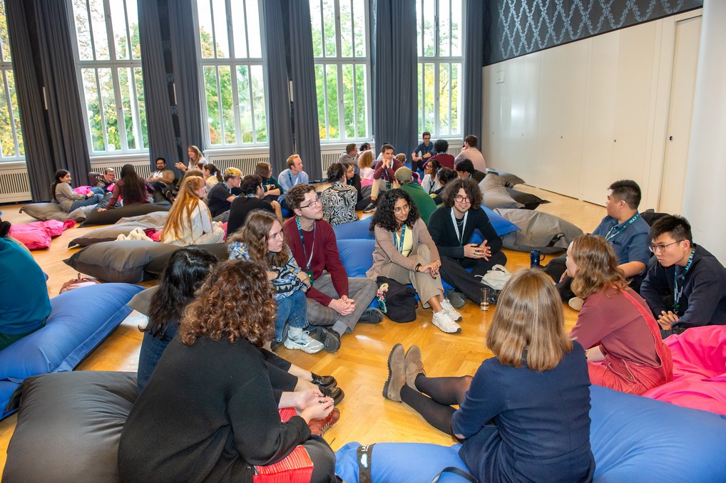 Mehrere Gruppen von Personen sitzen auf bunten Sitzsäcken in einem geräumigen Raum mit hohen Fenstern und hellen Holzböden.