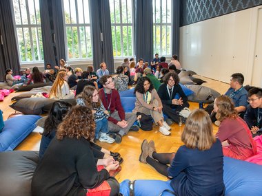 Multiple groups of people sitting on colorful bean bags in a spacious room with large windows, engaged in conversation and discussion.