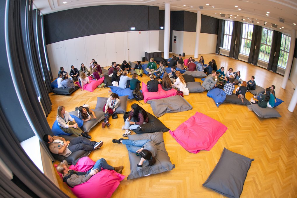 Viele Personen sitzen in einem großen Raum mit Holzfußboden auf bunten Sitzsäcken. Große Fenster lassen viel Tageslicht herein.