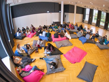 A large group of people sits on vibrant bean bags in a spacious room with wooden flooring and large windows.