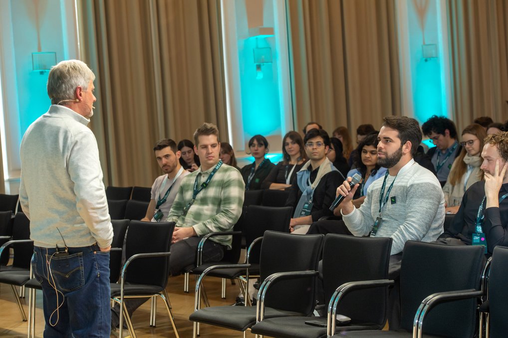 Ein Mann in einem grauen Pullover spricht in einem Konferenzraum zu sitzenden Teilnehmern. Einer der Teilnehmer hält ein Mikrofon, während die Gruppe aufmerksam zuhört. Im Hintergrund sind Vorhänge und blaue Beleuchtung zu sehen.