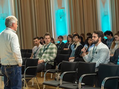 Ein Mann in einem grauen Pullover spricht in einem Konferenzraum zu sitzenden Teilnehmern. Einer der Teilnehmer hält ein Mikrofon, während die Gruppe aufmerksam zuhört. Im Hintergrund sind Vorhänge und blaue Beleuchtung zu sehen.