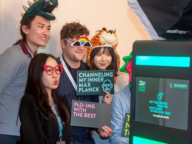 A group of individuals in playful hats, holding signs referencing Max Planck and PhD achievements, stand beside a photo booth display.