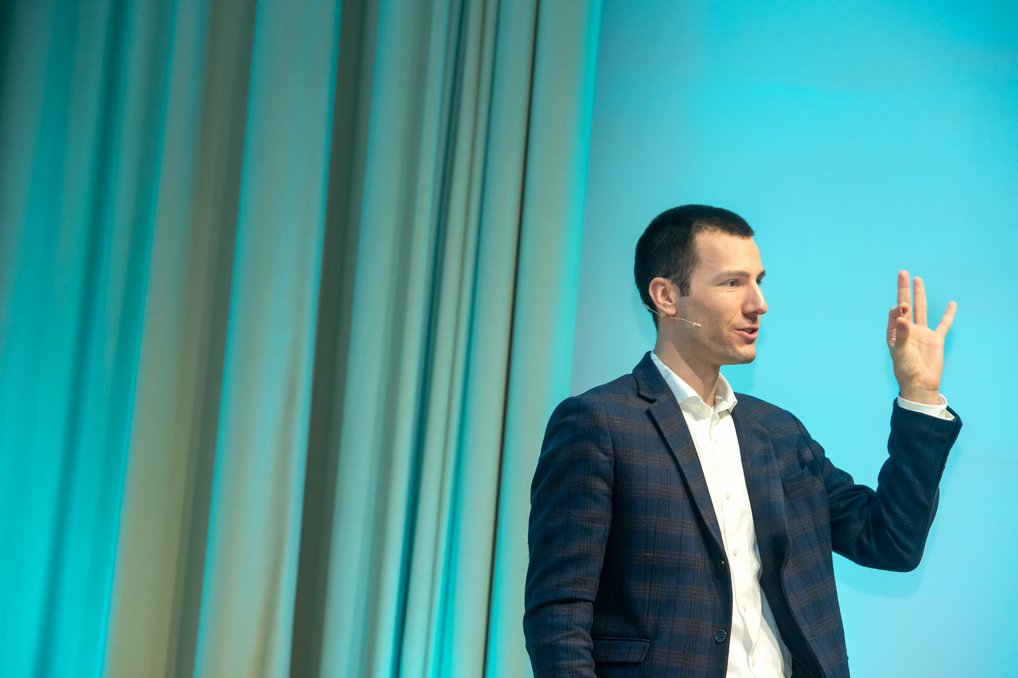 Eine Person in einem karierten Anzug steht vor einem blau beleuchteten Hintergrund und hebt die Hand mit gespreizten Fingern. Vorhänge sind im Hintergrund sichtbar.