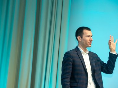 Eine Person in einem karierten Anzug steht vor einem blau beleuchteten Hintergrund und hebt die Hand mit gespreizten Fingern. Vorhänge sind im Hintergrund sichtbar.