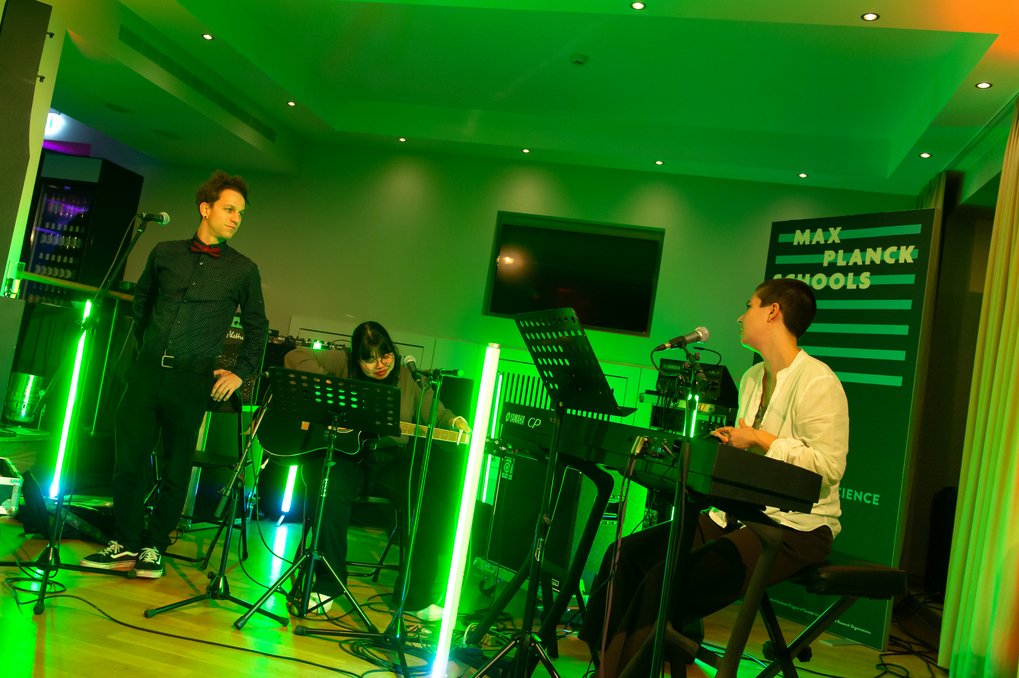 Drei Personen mit Mikrofon, Gitarre und Keyboard, umgeben von grünen Lichtern in einem Veranstaltungsraum.