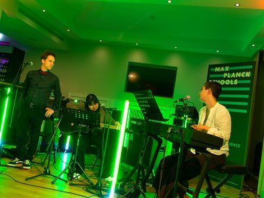 A group of musicians performing with a keyboard and guitar under green lighting. A sign for "Max Planck Schools" is displayed in the background.