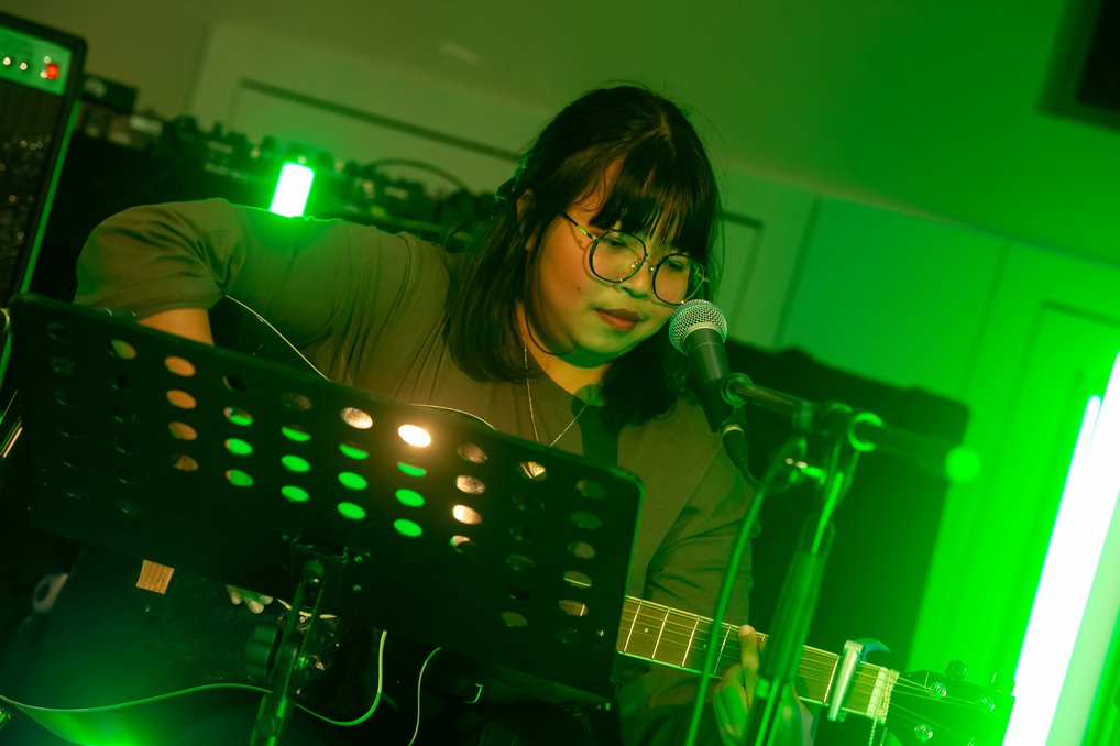 Eine Person spielt Akustikgitarre bei grünem Bühnenlicht, Mikrofon vorhanden, ein Notenständer hält Notenblätter.